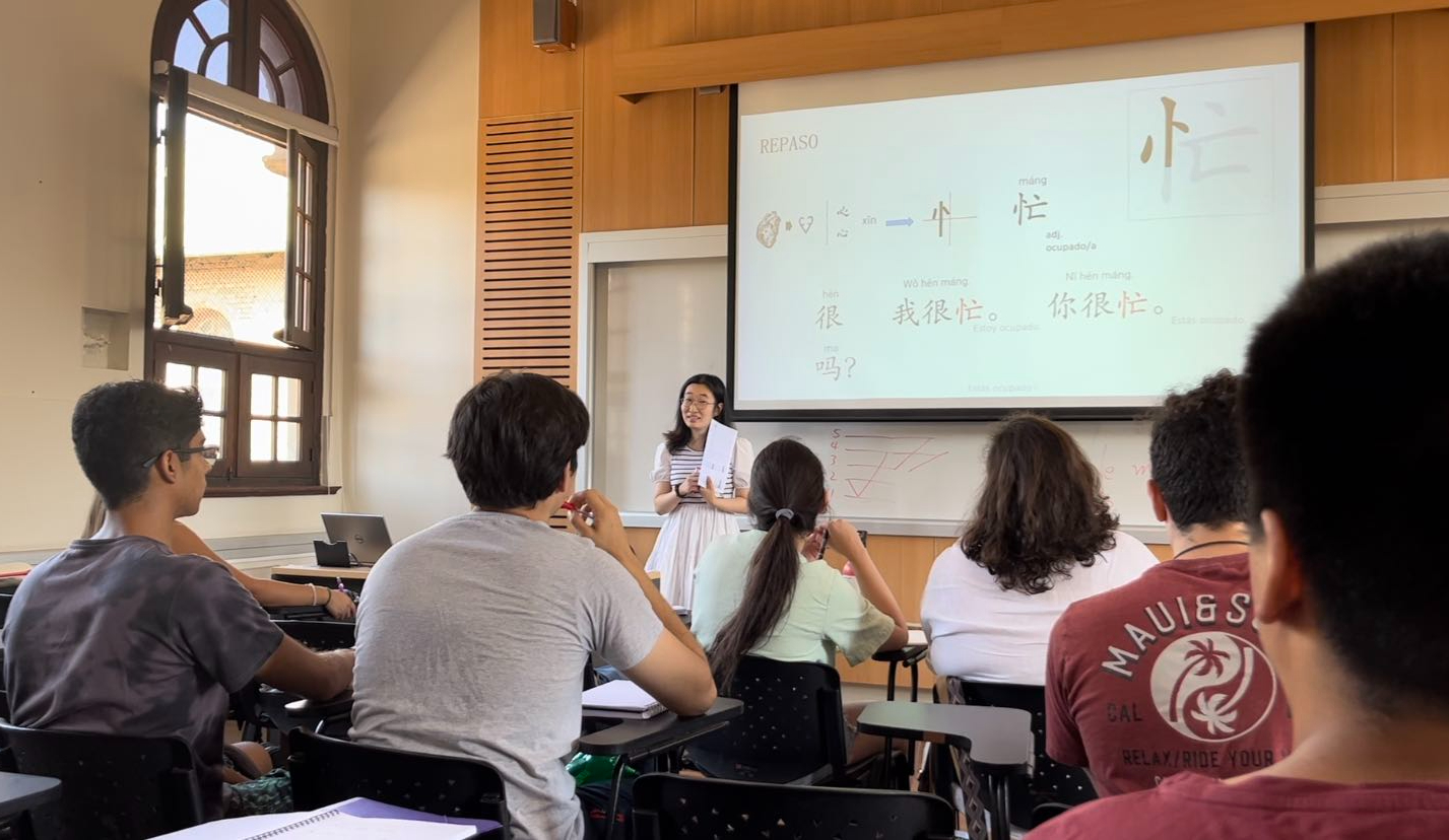 Profesora china enseñando chino a estudiantes en una sala, cuya pizarra exhibe caracteres chinos.