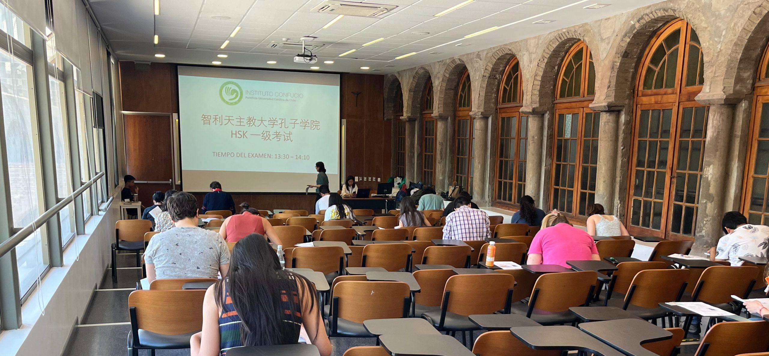 Varios alumnos rindiendo el examen de chino HSK en una sala grande, al fondo la pizarra exhibe la información.